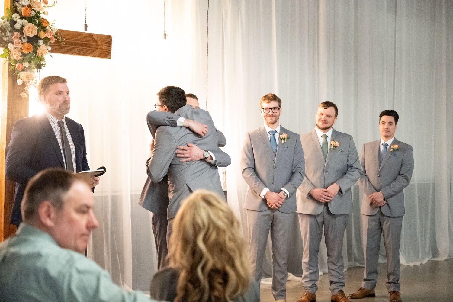 Groom embraces a groomsman at the altar while wedding party looks on during ceremony.