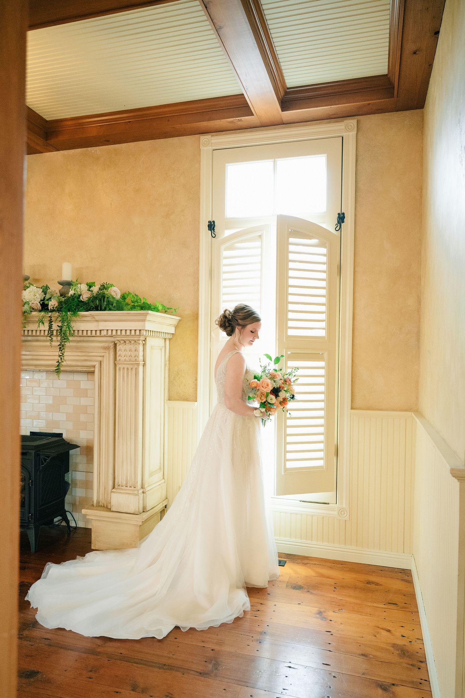 Bride in white gown holding bouquet stands by shuttered window near fireplace on hardwood floors.