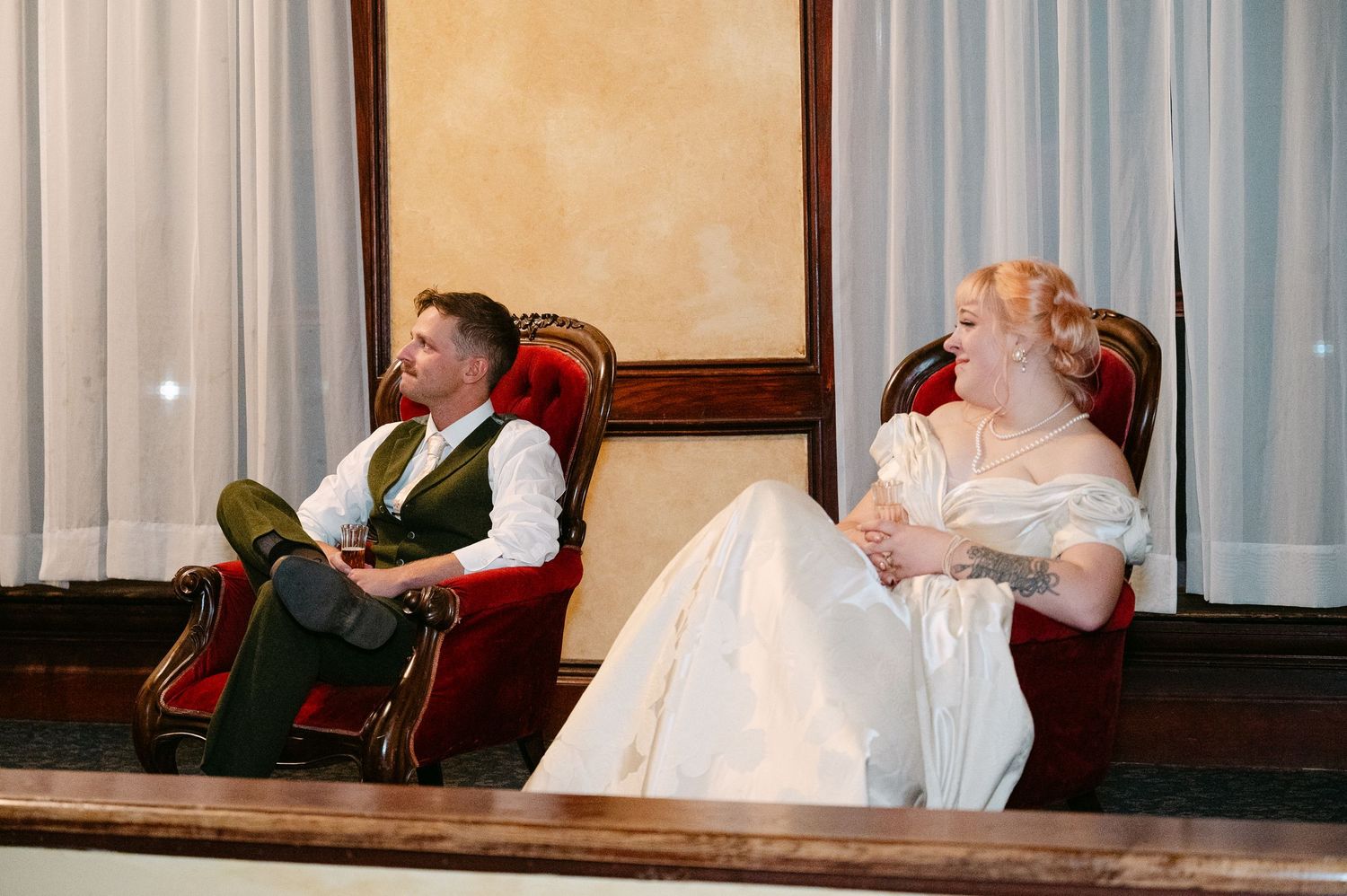 Central MN Wedding Photos | Bride in champagne gown and groom in olive suit relaxing in red velvet chairs during wedding reception.