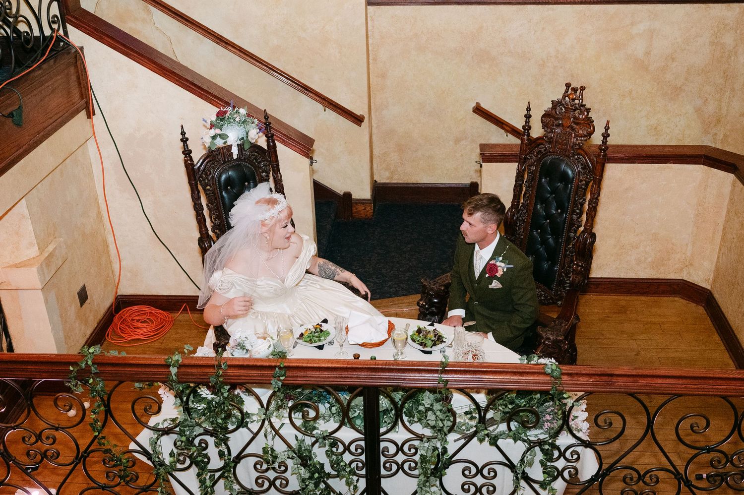 Central MN Wedding Photos | Bride and groom sit in ornate throne chairs at sweetheart table on elegant balcony during reception.