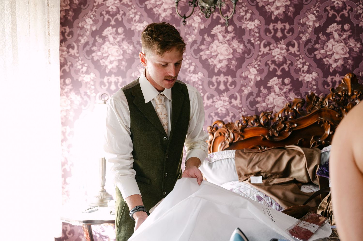 Central MN Wedding Photos | Groom in white shirt and dark vest carefully handling wedding dress on bed before ceremony.