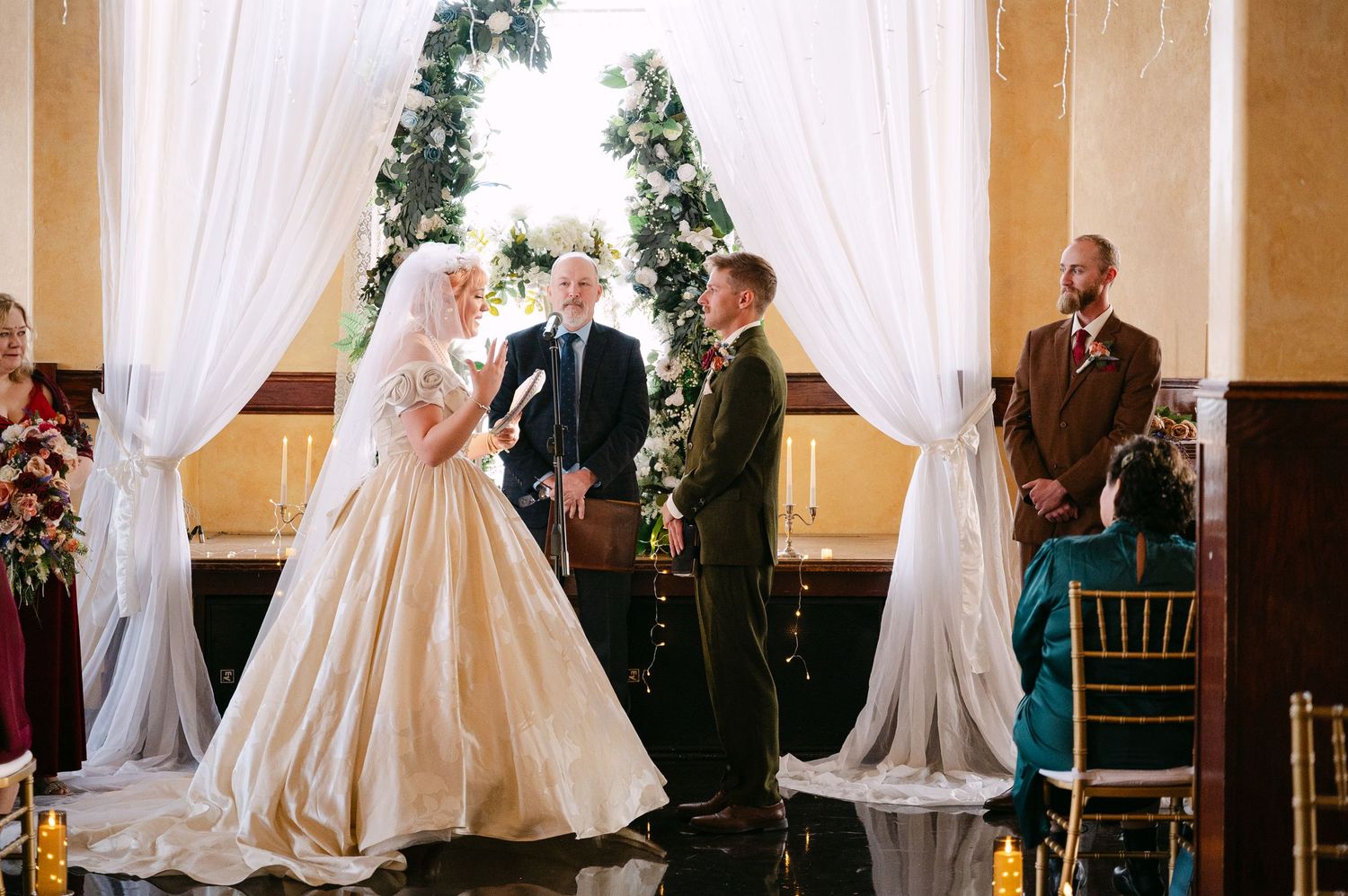 Central MN Wedding Photos | Bride and groom exchanging vows at altar with white draping and Christmas tree in elegant ballroom.