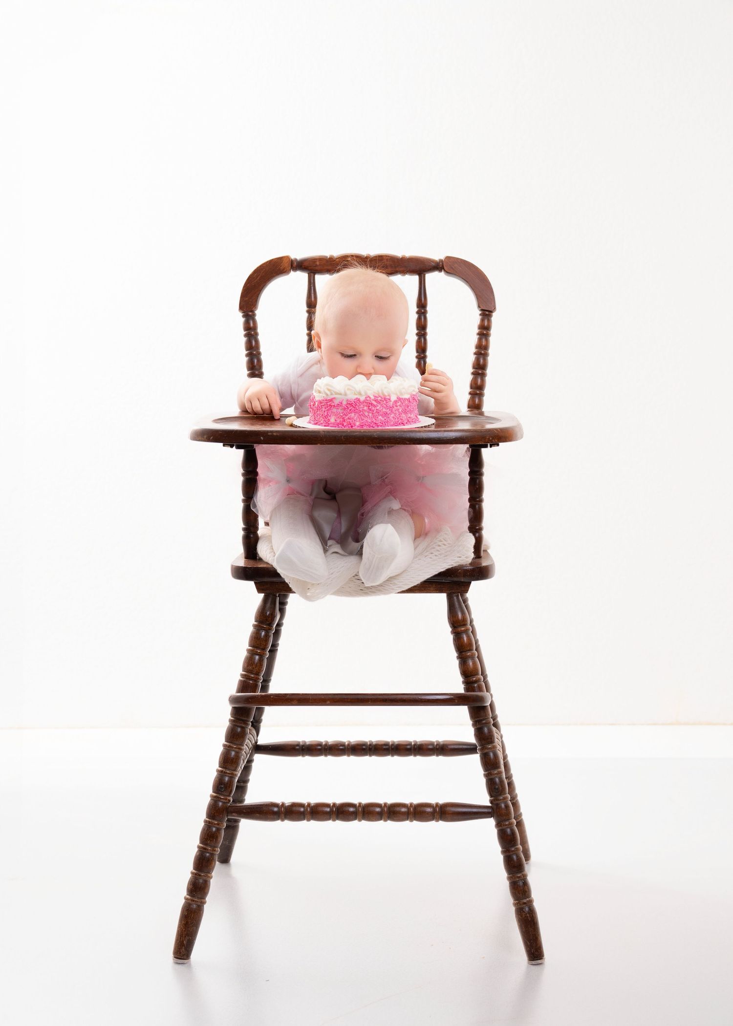 Central MN Cake Smash Photography - Baby in pink tutu sitting in wooden high chair eating a pink birthday cake on white background.