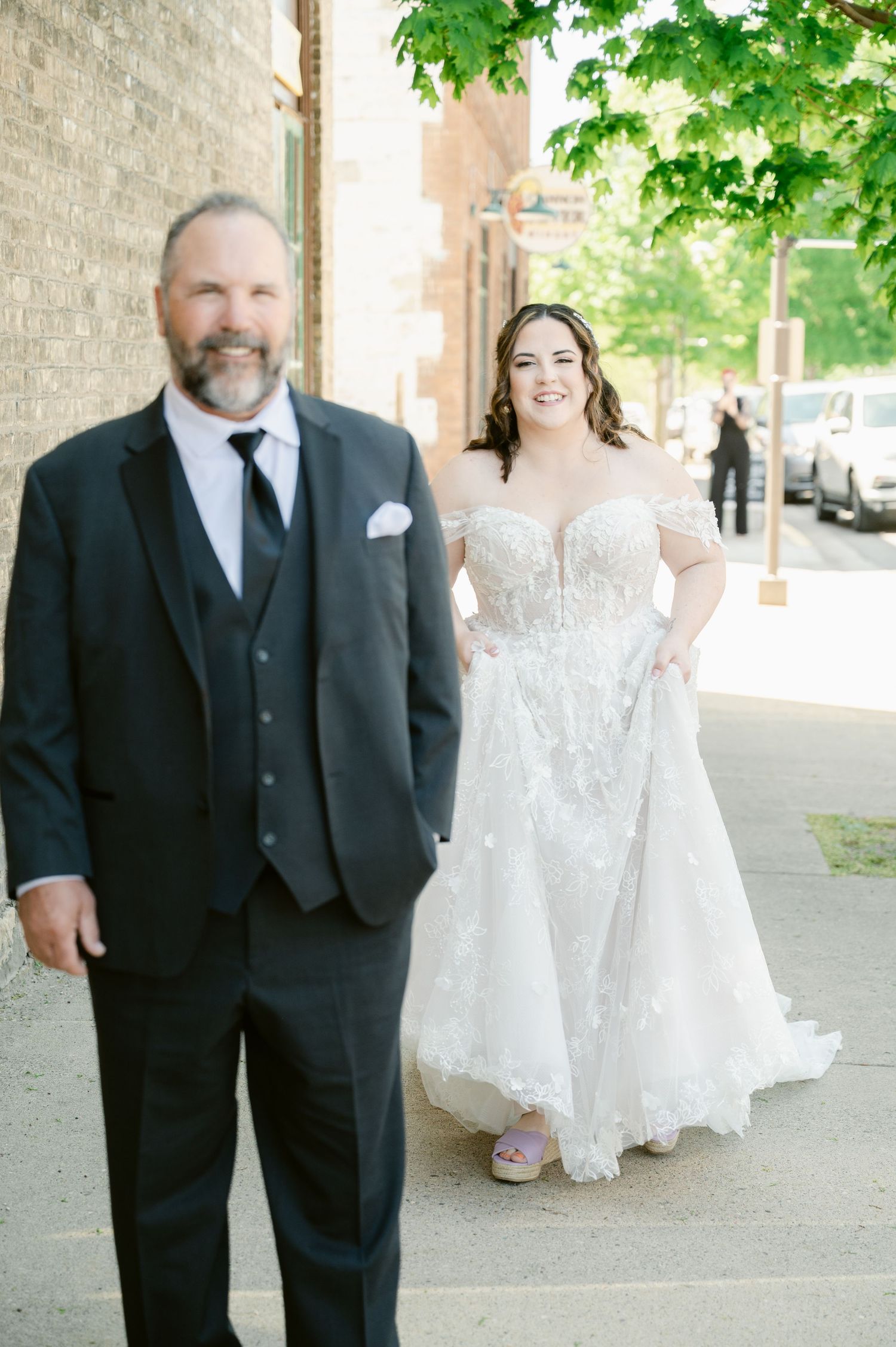 Central MN Wedding Photos - Bride in floral lace off-shoulder wedding gown walks with her father on a sunny sidewalk.