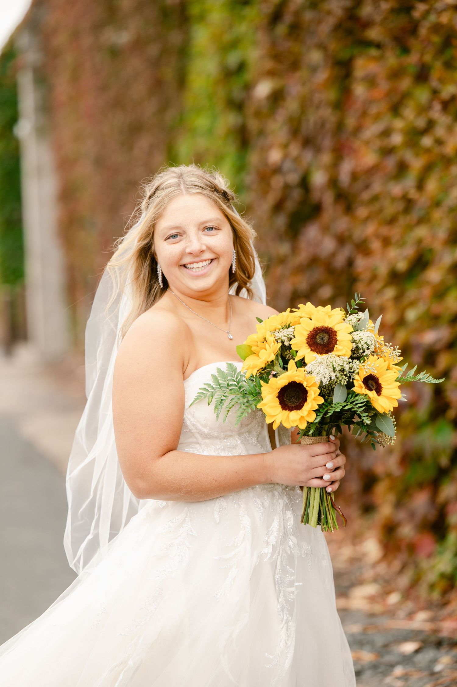 Central MN Wedding Photos | Smiling bride in strapless lace gown holding vibrant sunflower bouquet against ivy wall.