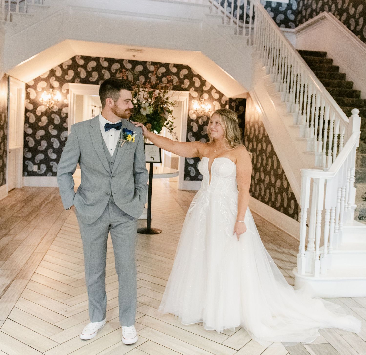 Central MN Wedding Photos | Bride in strapless tulle gown adjusts groom's gray suit lapel near elegant staircase with floral wallpaper.