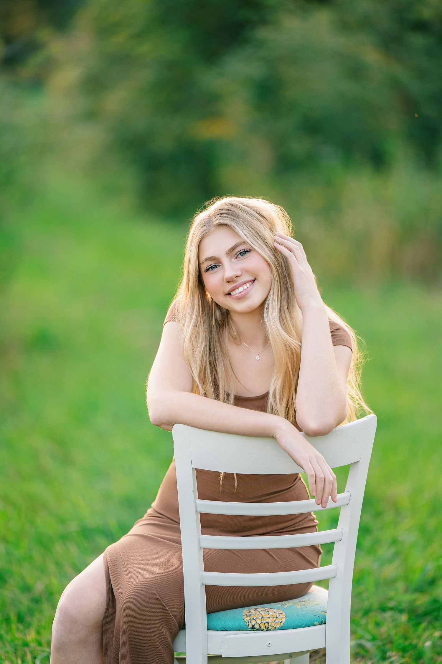 Central MN Senior Photography - Smiling blonde woman in brown dress sitting on white chair outdoors in green field during golden hour.