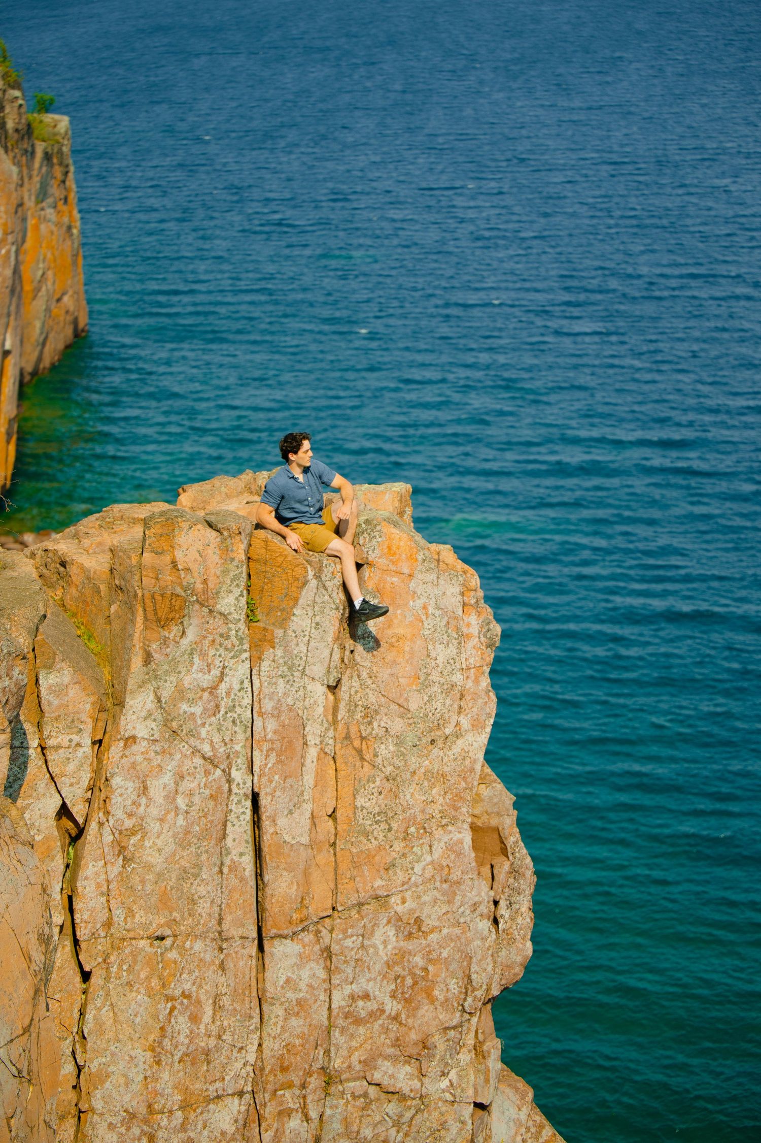 A man sits on a rocky cliff edge overlooking turquoise ocean waters on a sunny day.