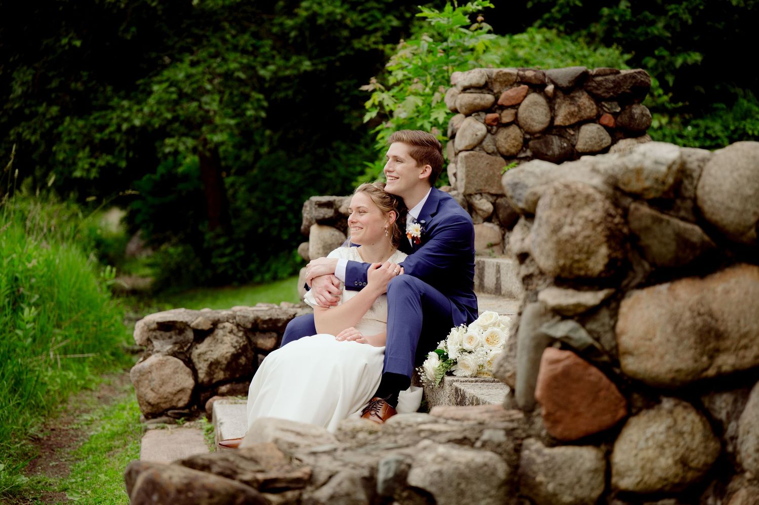 A couple in wedding attire share a romantic moment while sitting on a rustic stone wall surrounded by lush greenery.