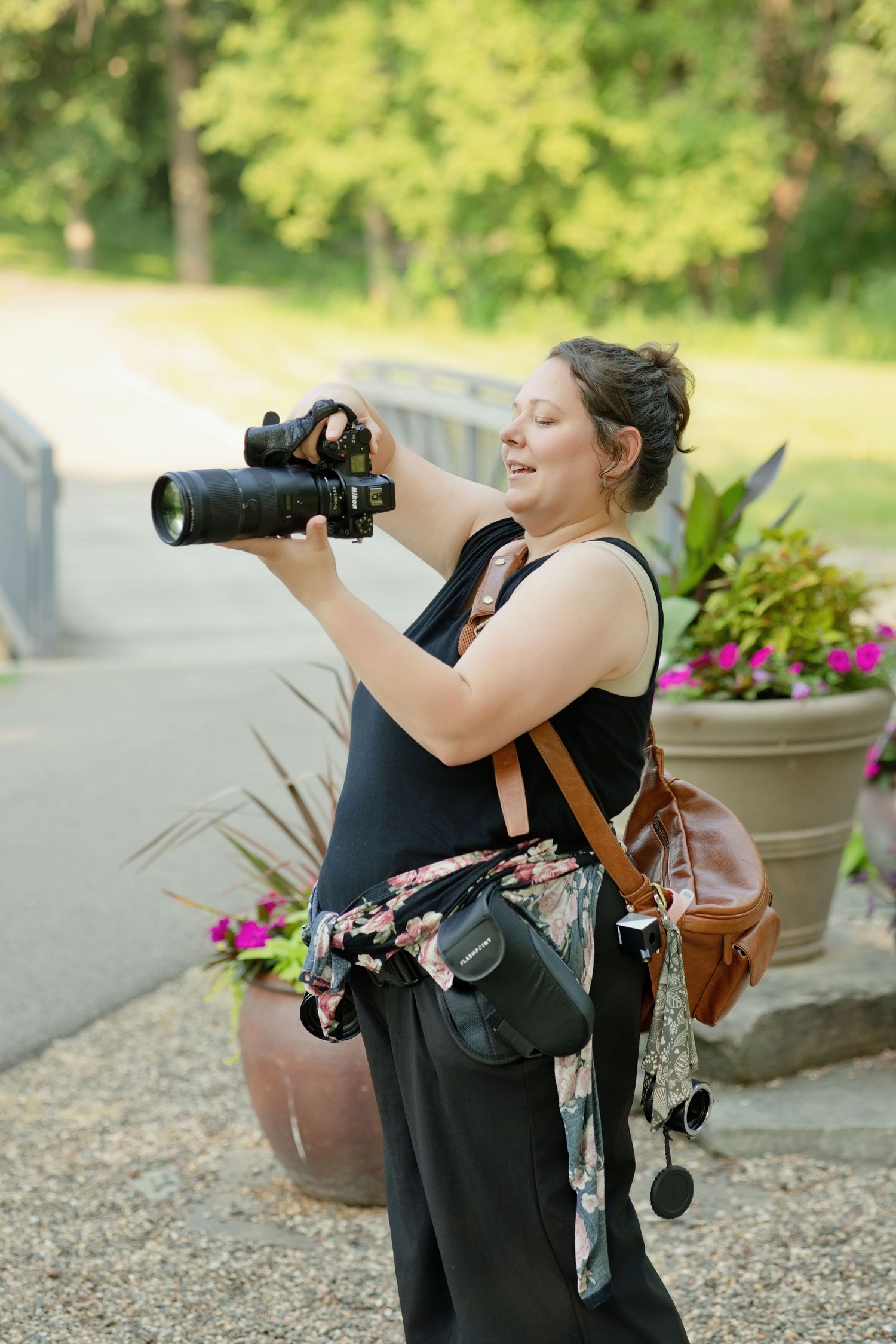 Woman photographer holding DSLR camera outdoors near colorful flower pots, wearing black outfit with brown leather bag.