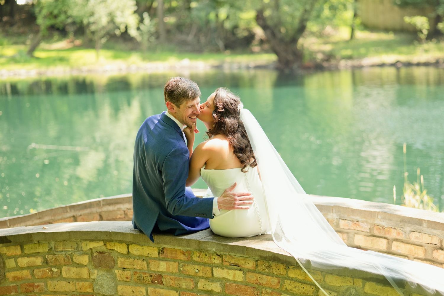 Bride and groom share a romantic moment sitting on a stone bridge over a serene turquoise lake on their wedding day.