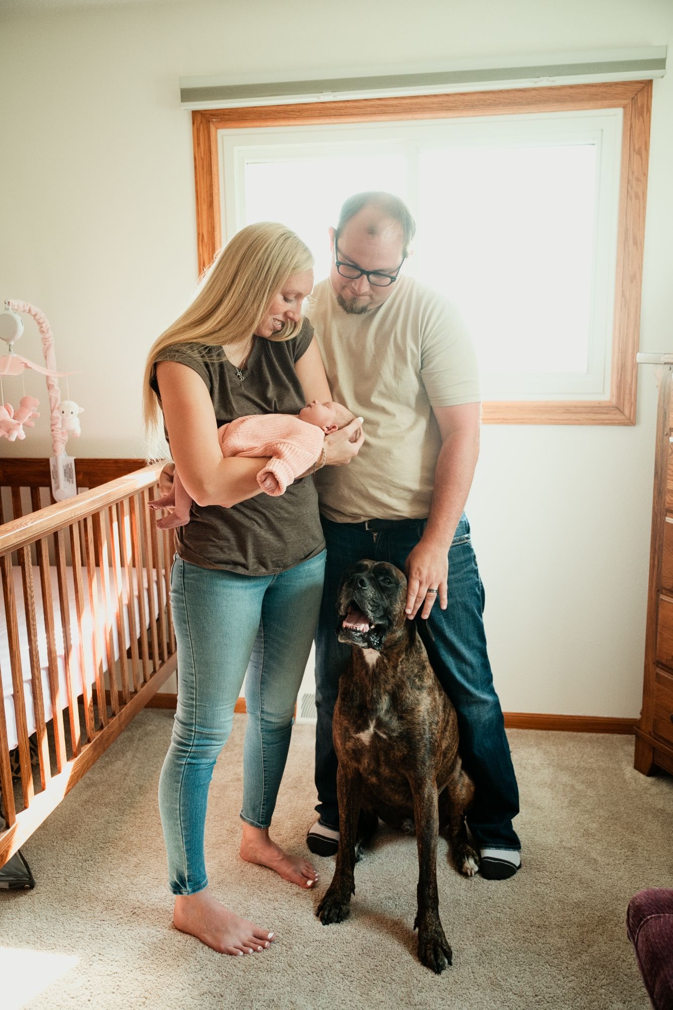 A couple with a newborn baby and a brindle dog standing in a nursery room next to a wooden crib.