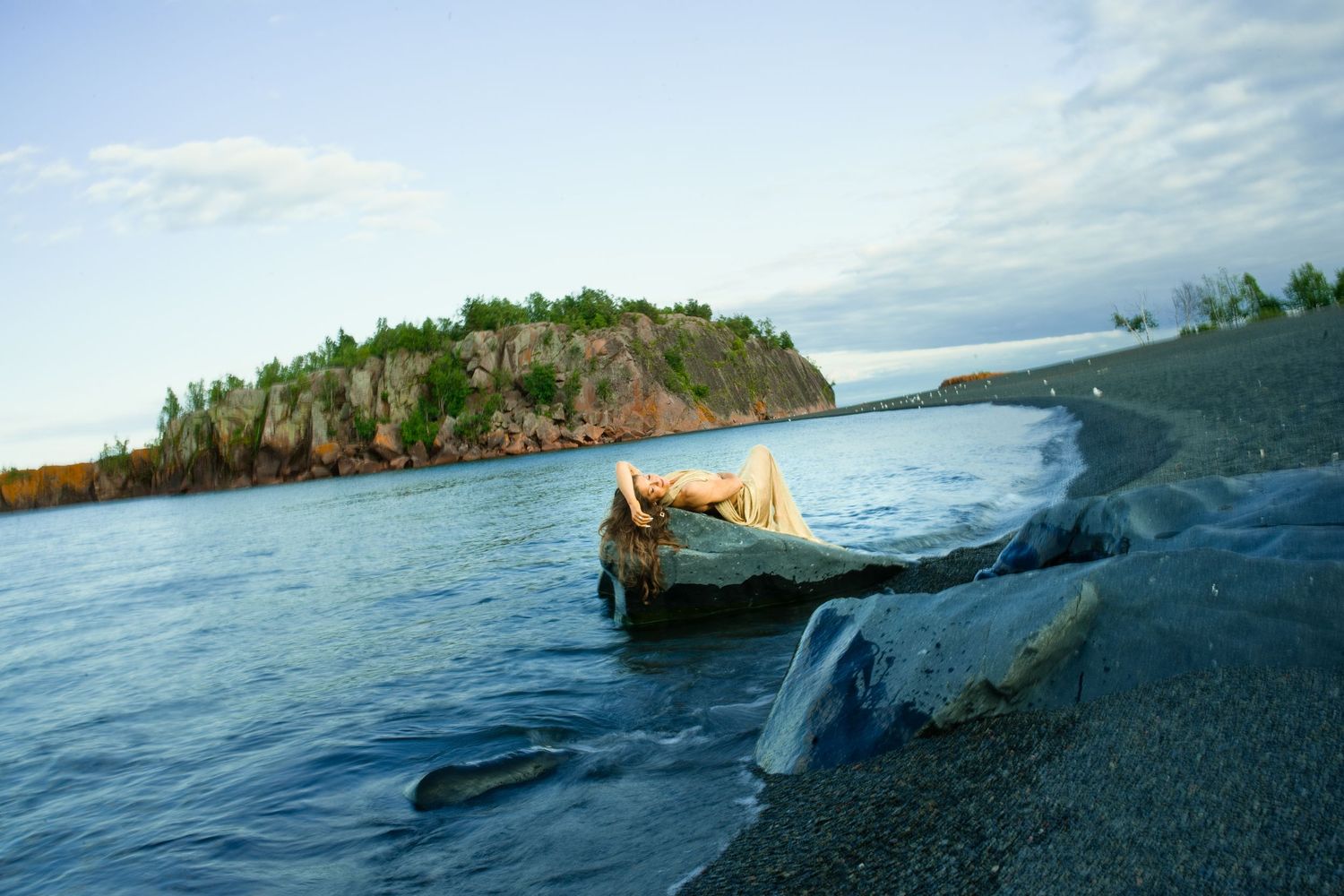 Person lying on a large rock at the edge of a calm blue lake with a rocky tree-covered island and pebble shoreline under a soft sky.