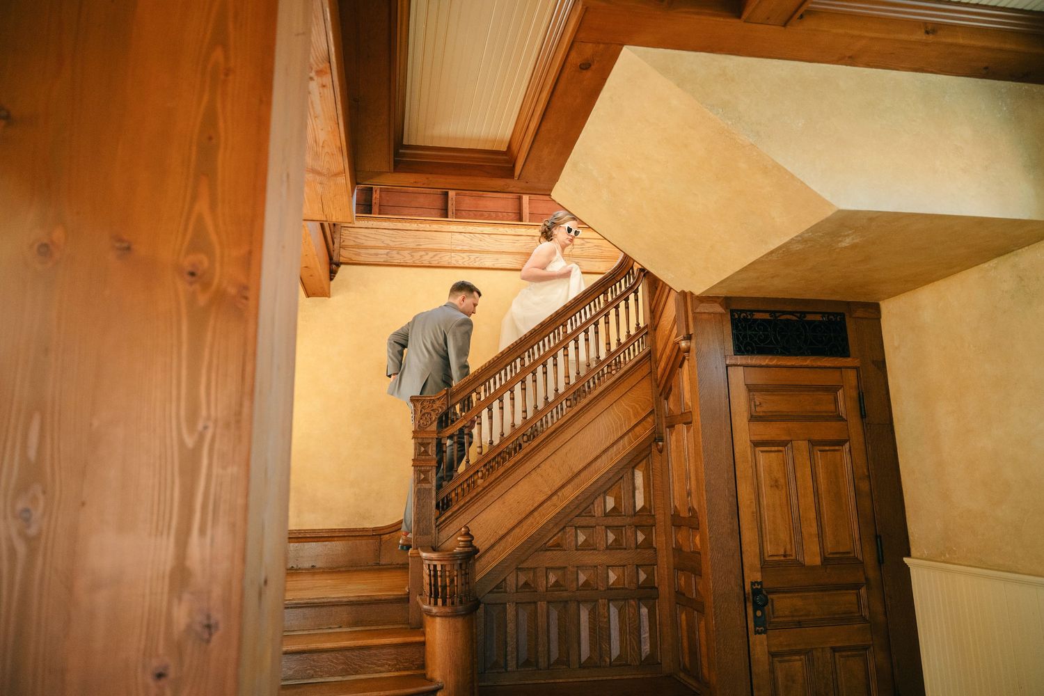 Bride and groom share a romantic moment on elegant wooden staircase in a grand historic venue.