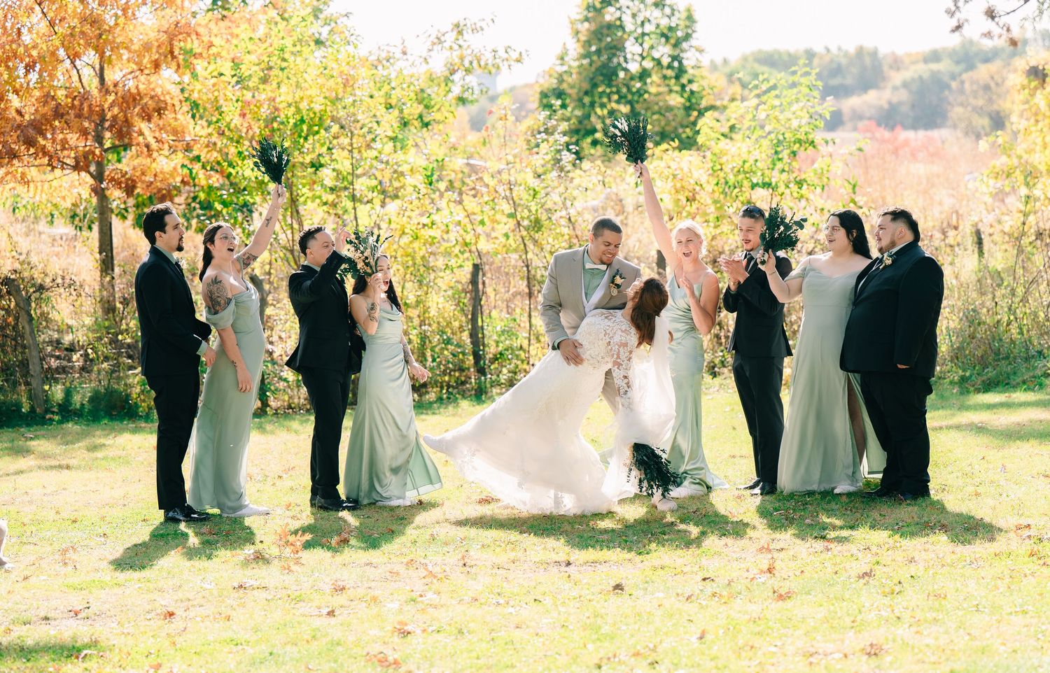 Wedding party celebrates outdoors in autumn foliage, with bride and groom embracing at center.