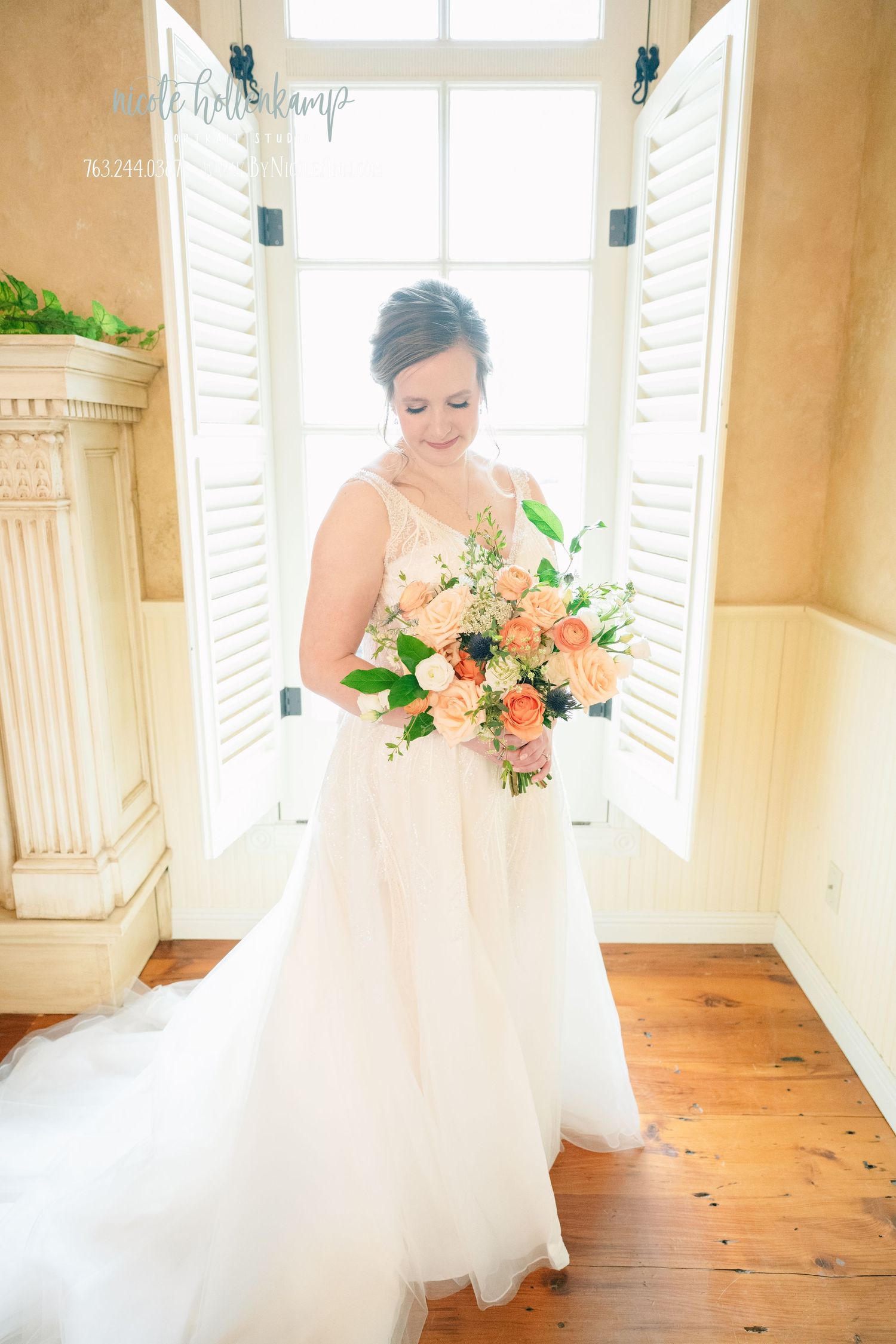 A bride in a flowing white gown holds a peach and coral bouquet while standing in a sunlit room with wooden floors.
