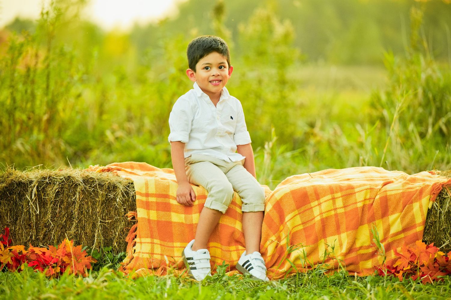 A smiling boy sitting on a hay bale with an orange blanket in a sunny field.