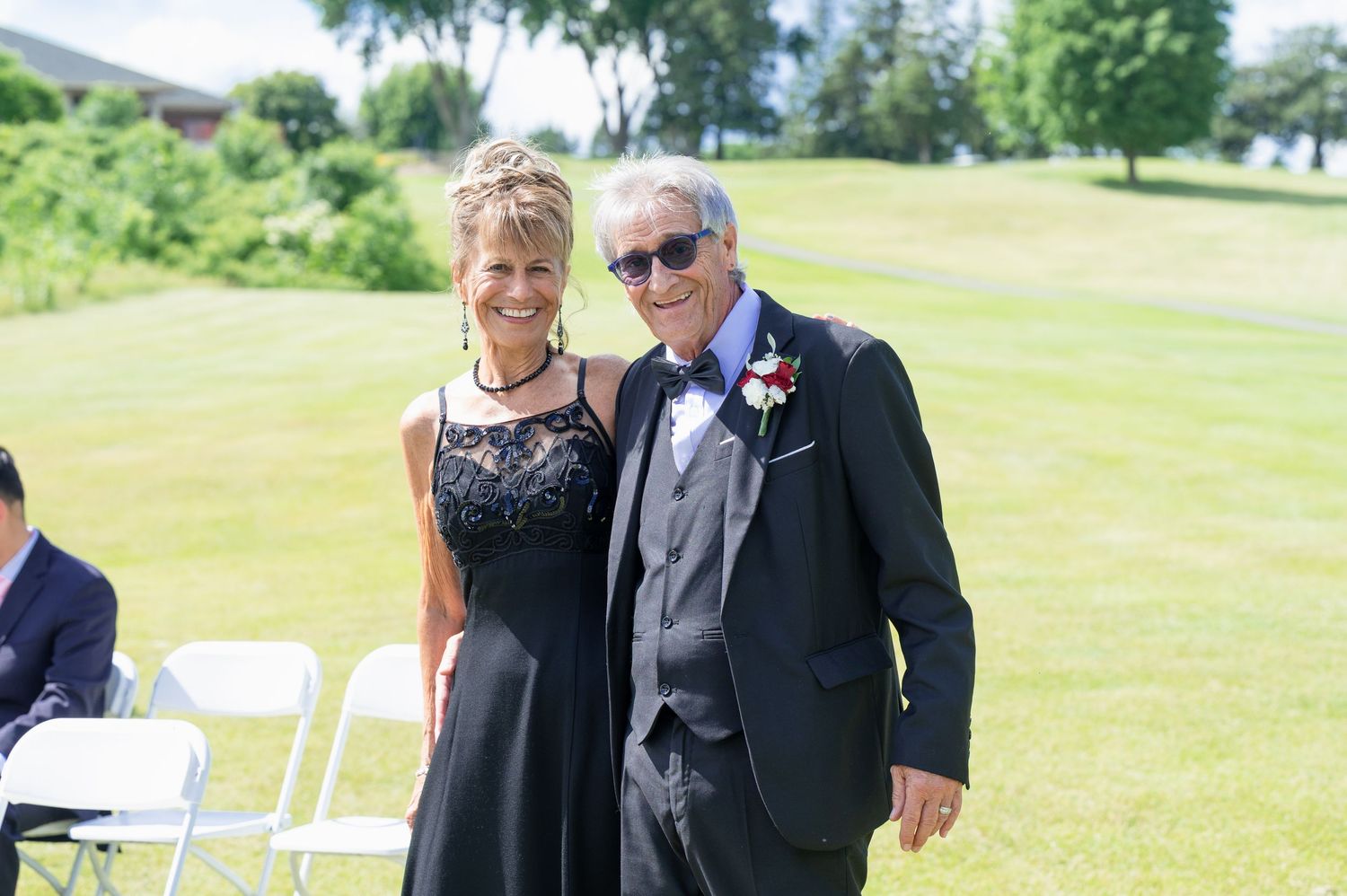 Smiling wedding guests, woman in black lace dress and man in suit with boutonniere, pose outdoors.