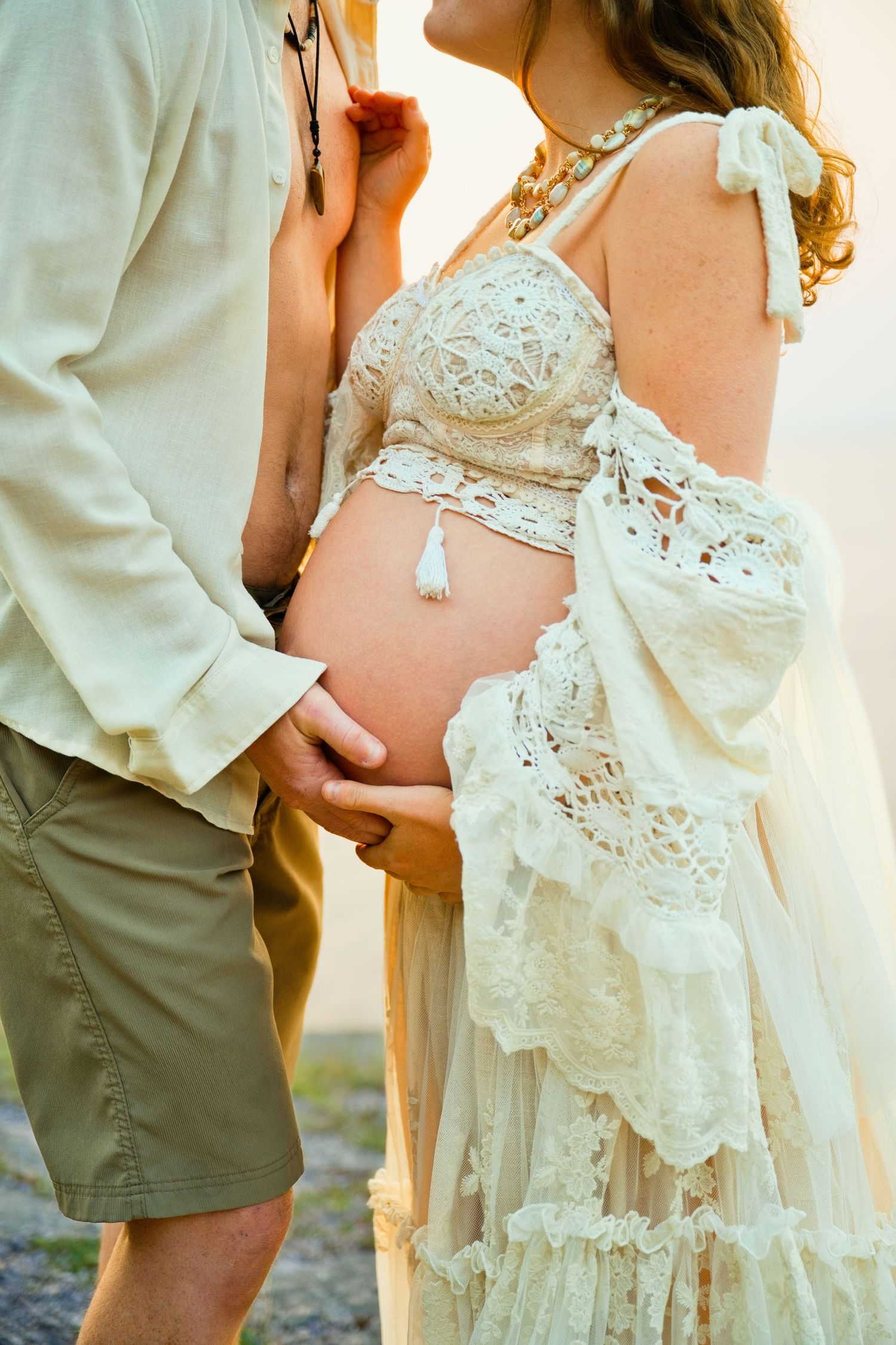 Couple cradling pregnant belly outdoors, woman in boho lace dress, golden hour maternity photo session.