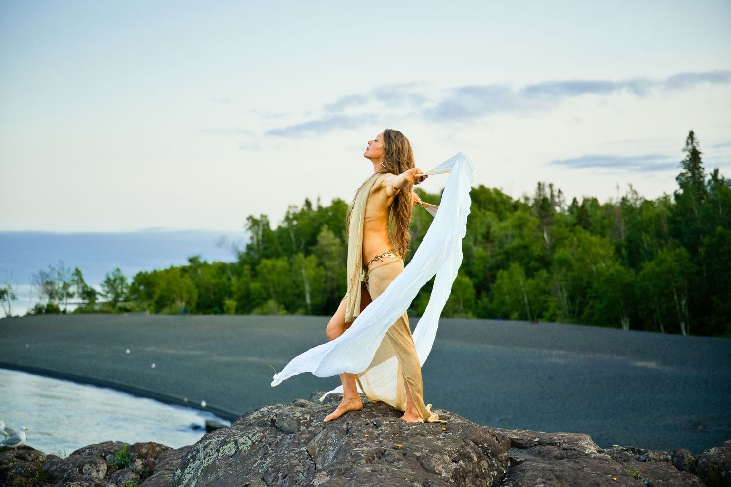 Woman in gold dress holding white fabric on rocky shore with forest and water backdrop.
