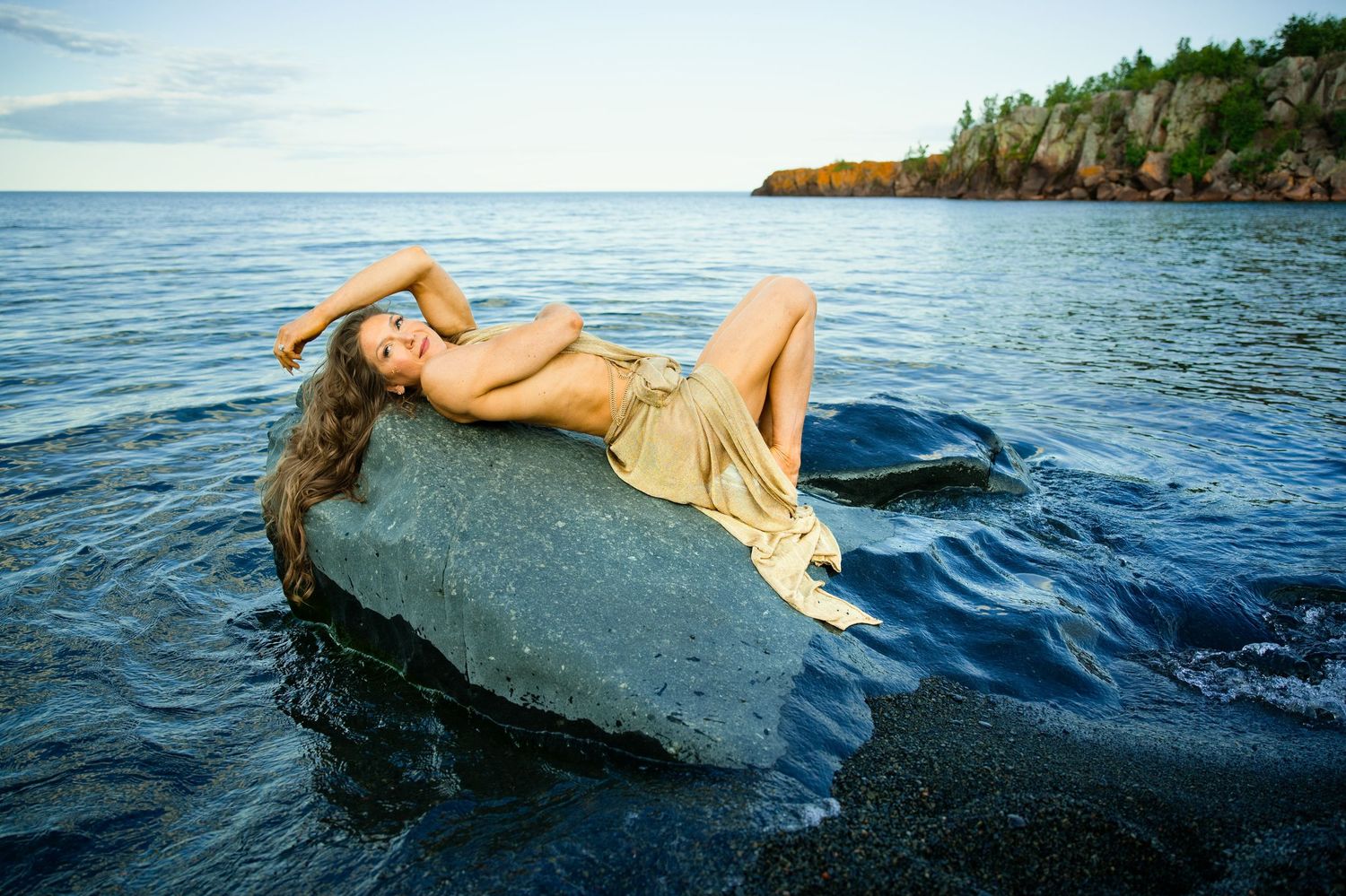 Woman in gold dress reclining on a large rock surrounded by calm ocean waters with a rocky coastline in the background.
