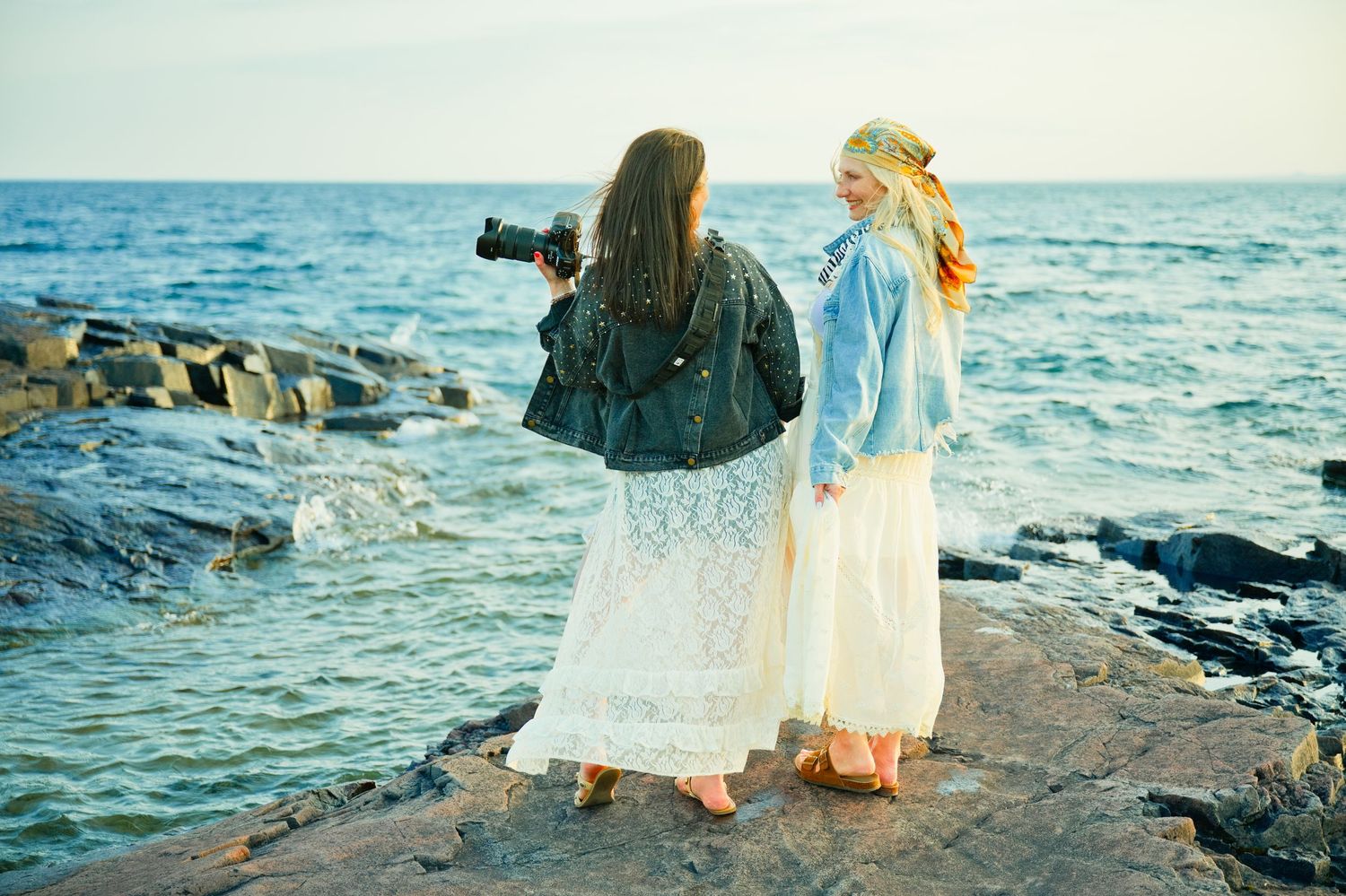 Two women in white lace skirts and denim jackets stand on rocky shore, one photographing the ocean.