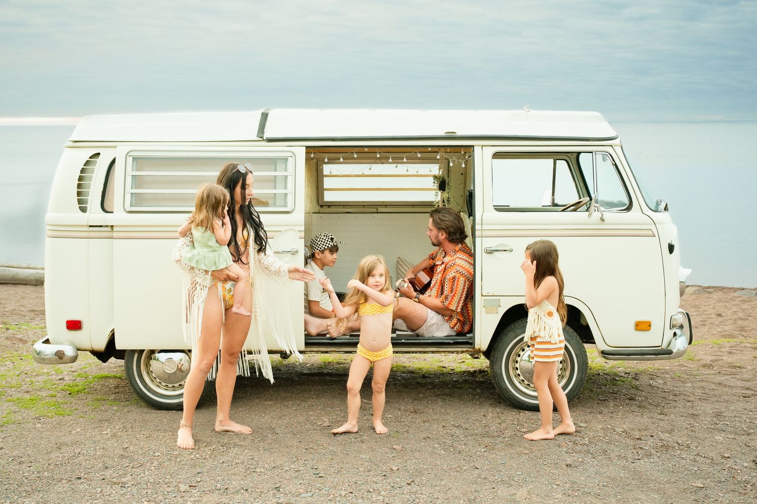 Family with children gathered around a classic VW camper van parked on a beach shoreline.