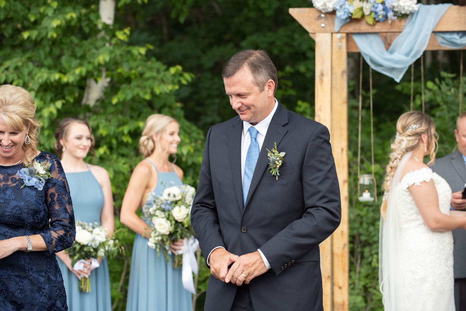 Outdoor wedding ceremony under wooden arbor with floral decorations and light blue bridesmaid dresses. Zimmerman MN Weddings