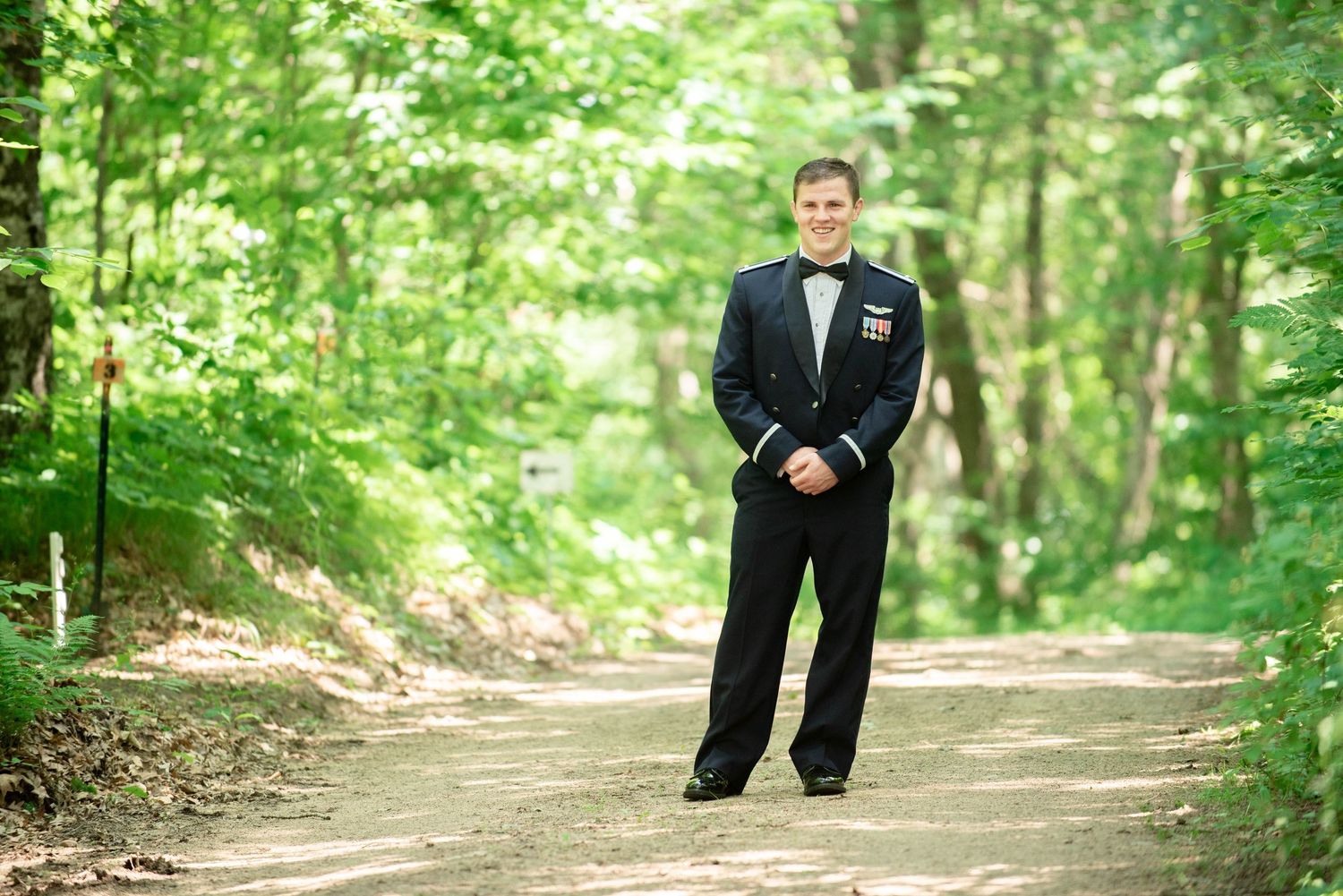 A groom in formal military attire stands on a wooded path. Zimmerman MN Weddings