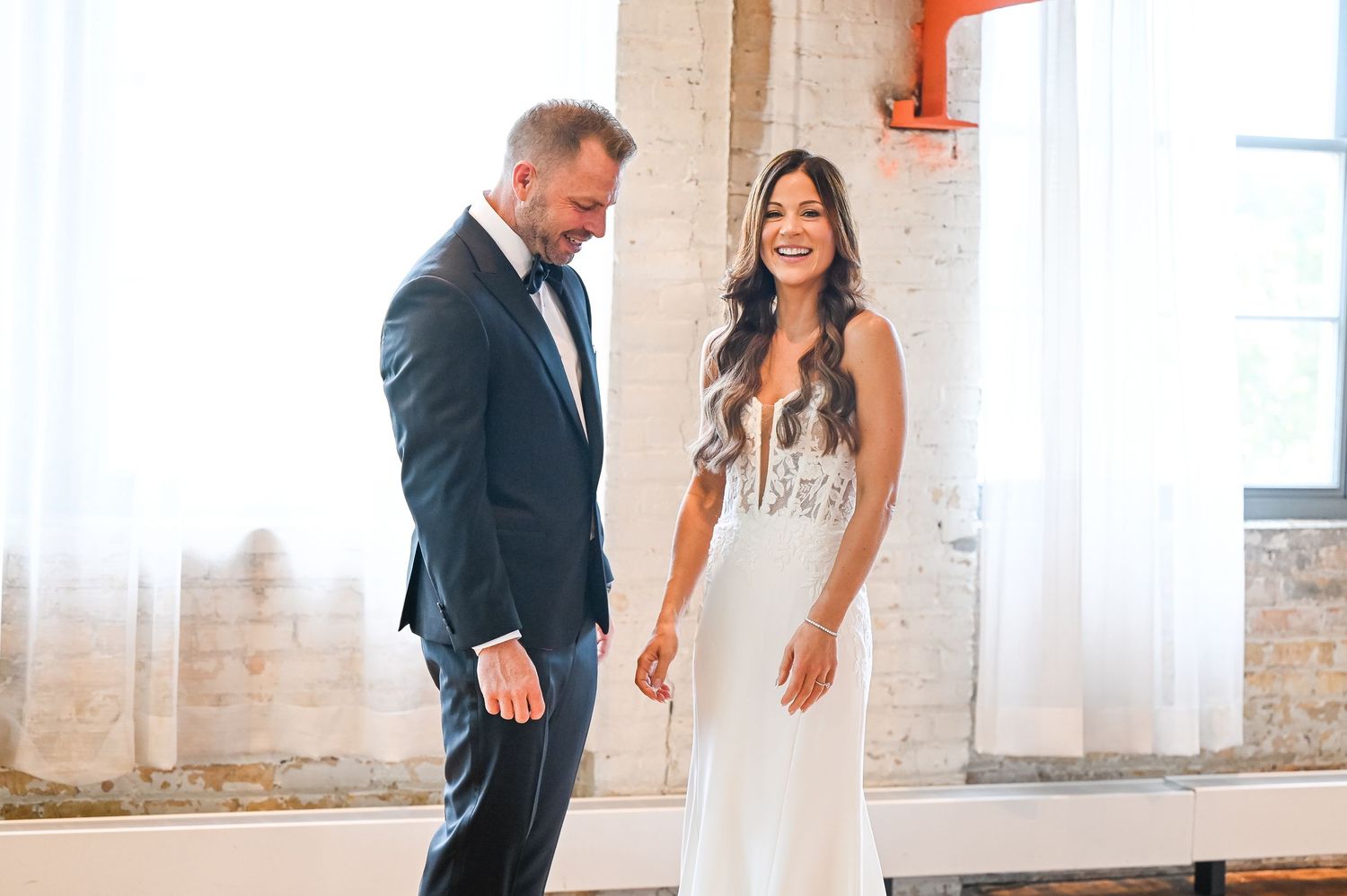 A couple interacts playfully in a sunlit venue with exposed brick walls and white curtains.