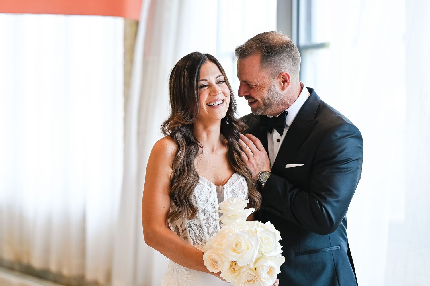Joyful wedding couple shares intimate moment while holding white rose bouquet.