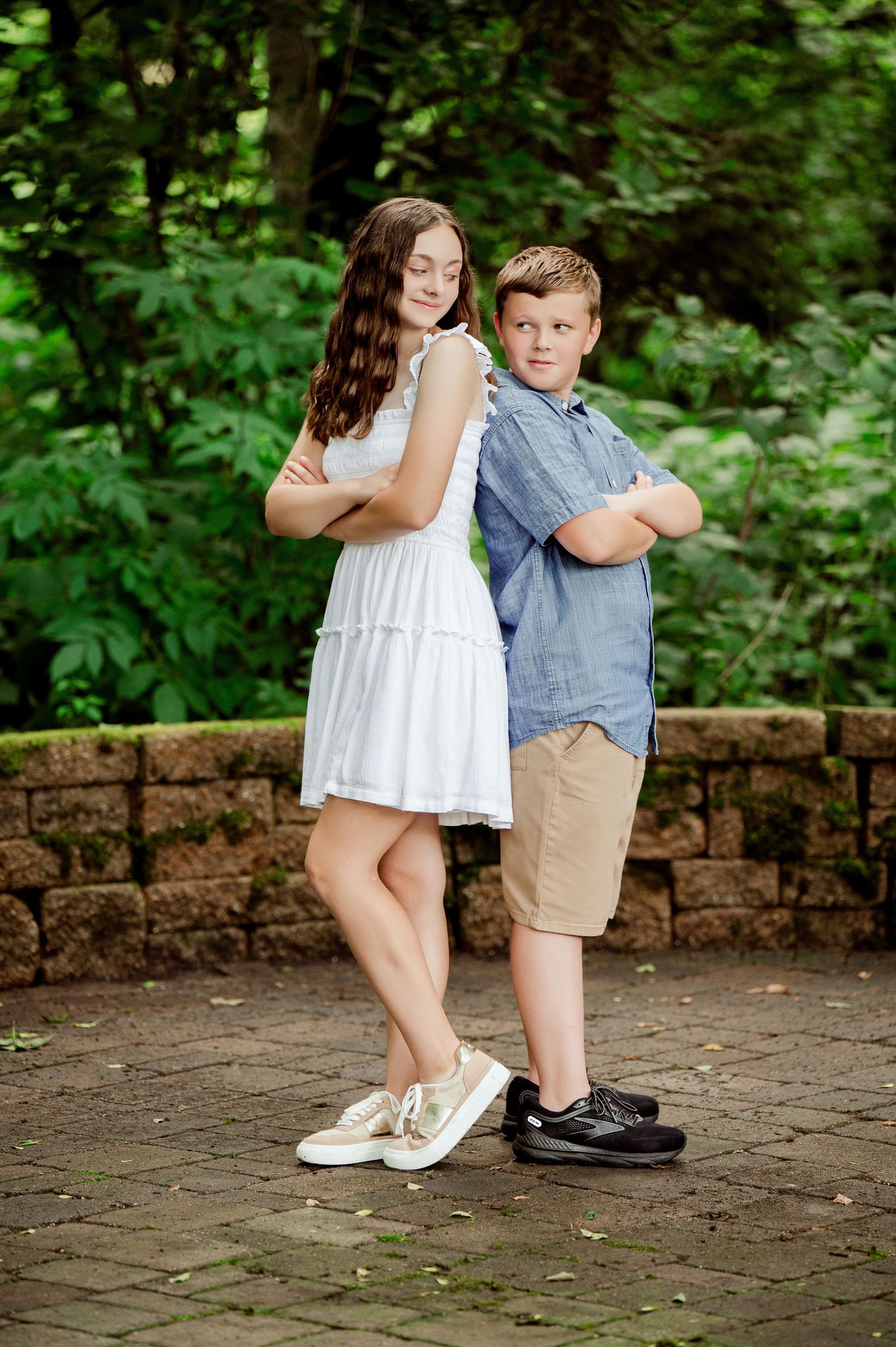 Two siblings pose back-to-back on a stone patio with forest greenery in the background.