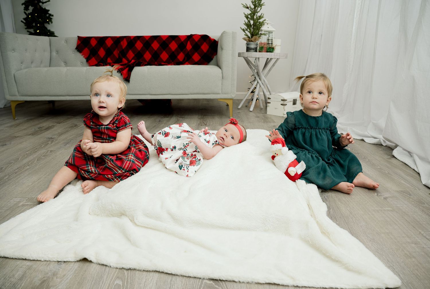 Two small children sit on a white blanket wearing holiday-themed buffalo plaid and green velvet outfits.