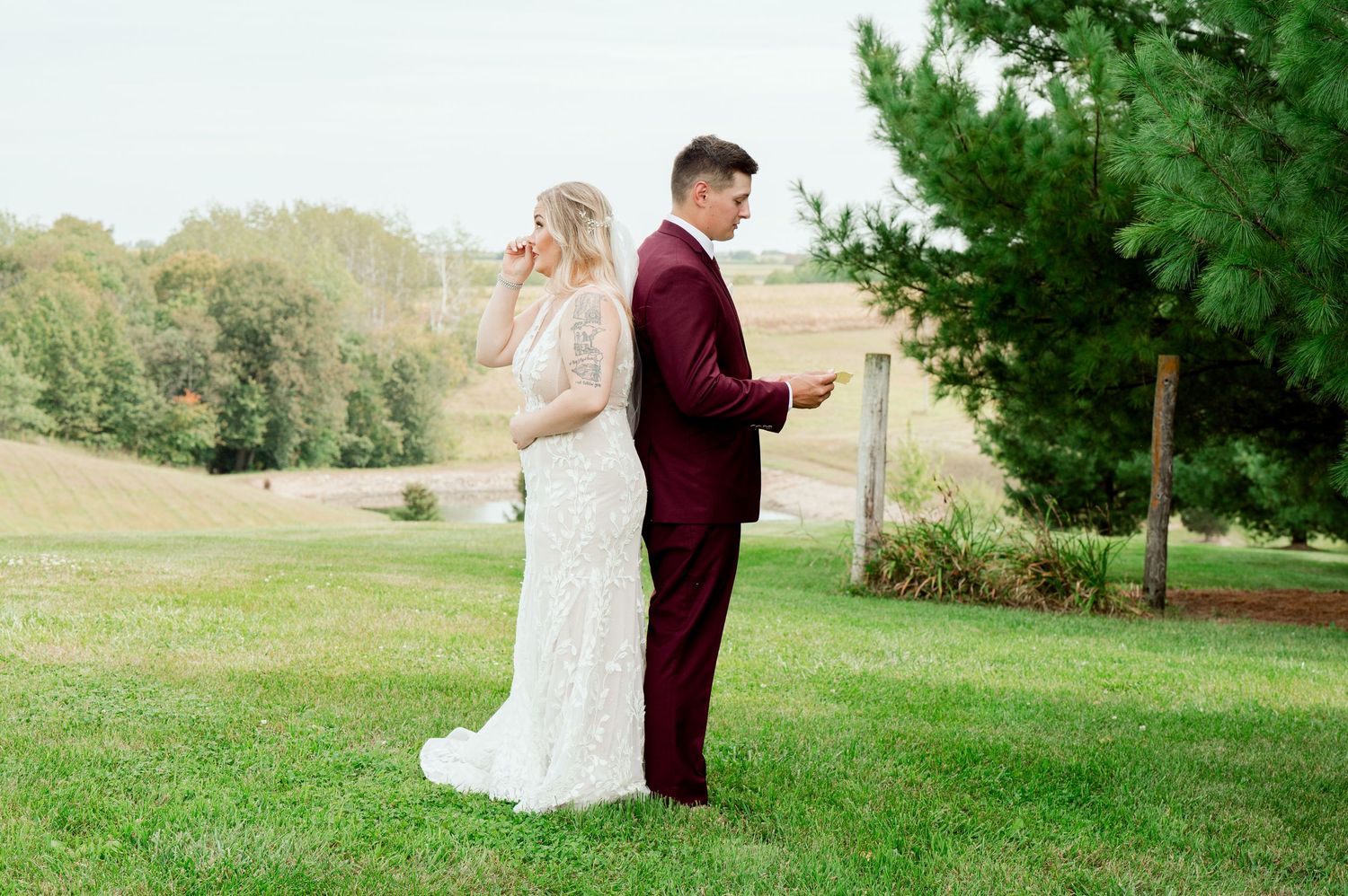 Wedding couple stands back-to-back on a grassy hill while reading private letters before their ceremony.
