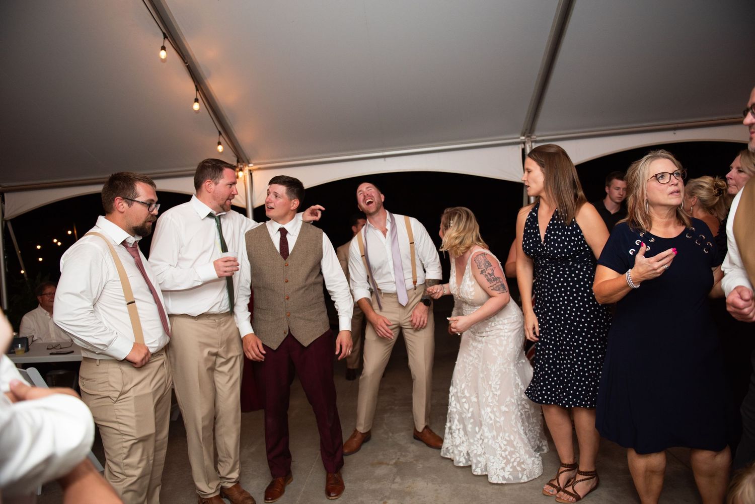 Wedding guests dancing and celebrating in a dimly lit reception tent with festive lighting and party decor.