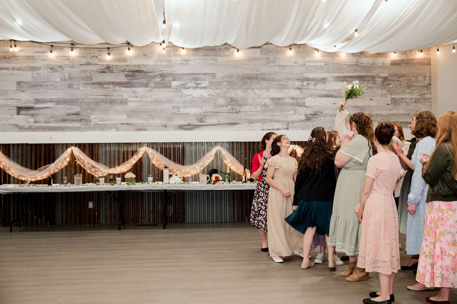 Wedding reception venue with rustic wood walls, white draping, and string lights decorating the head table area.