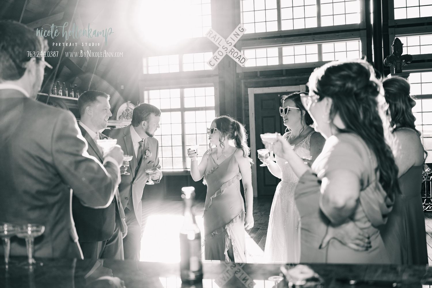 Black and white wedding reception photo with guests toasting drinks indoors near large windows with natural light. Crooked Willow Osakis