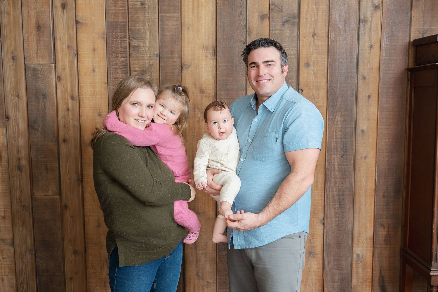 Family portrait of four people standing together against a rustic wooden wall background.. Minnesota milestone photographer