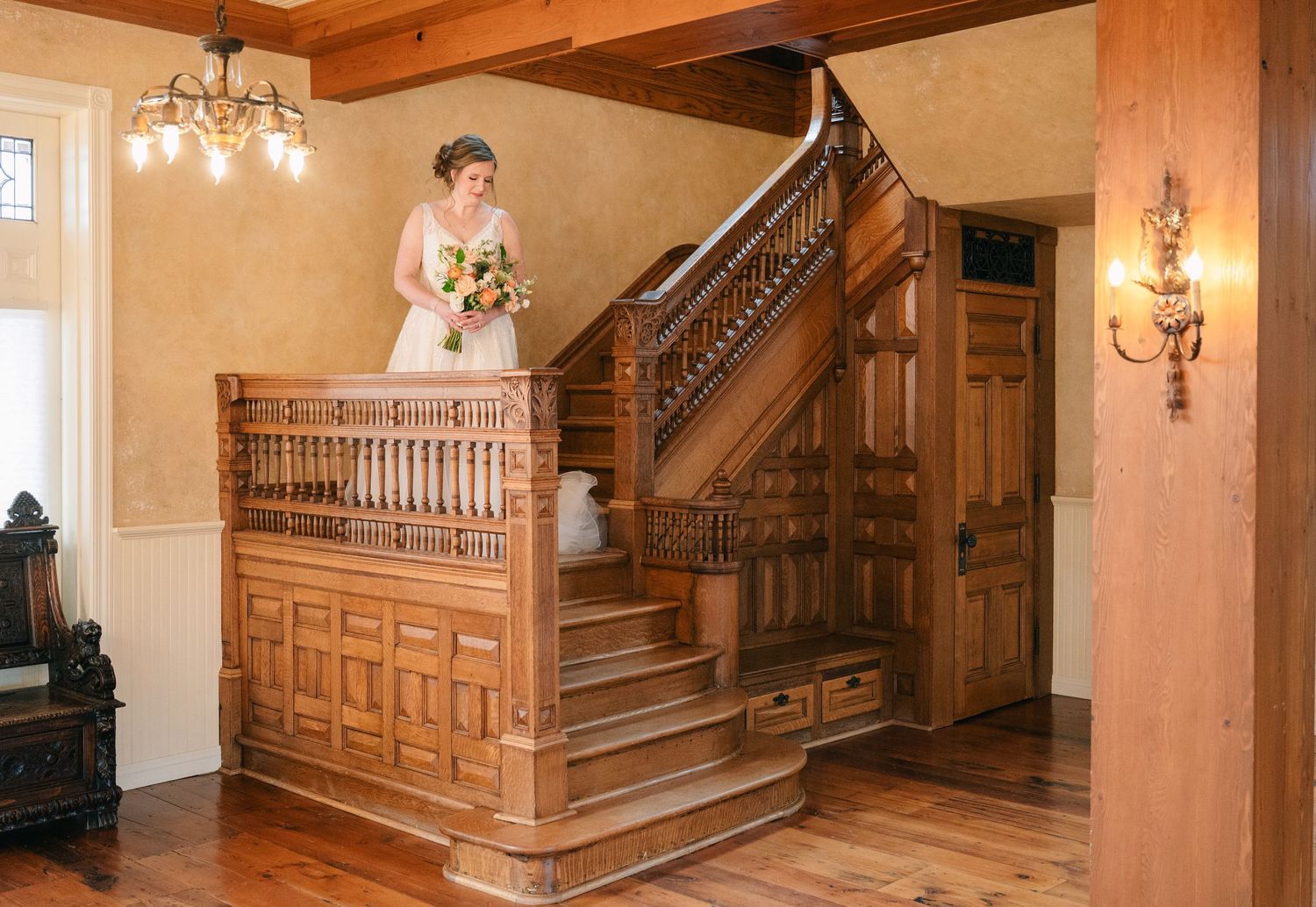 Bride in white gown with bouquet poses at top of ornate wooden staircase in historic venue.