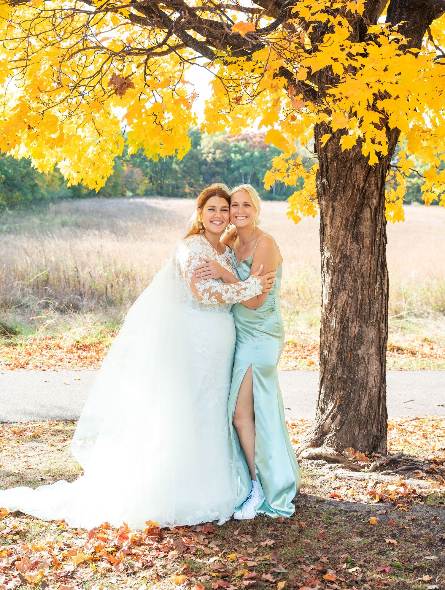Bride in white lace gown hugs bridesmaid in mint dress under vibrant yellow autumn tree.