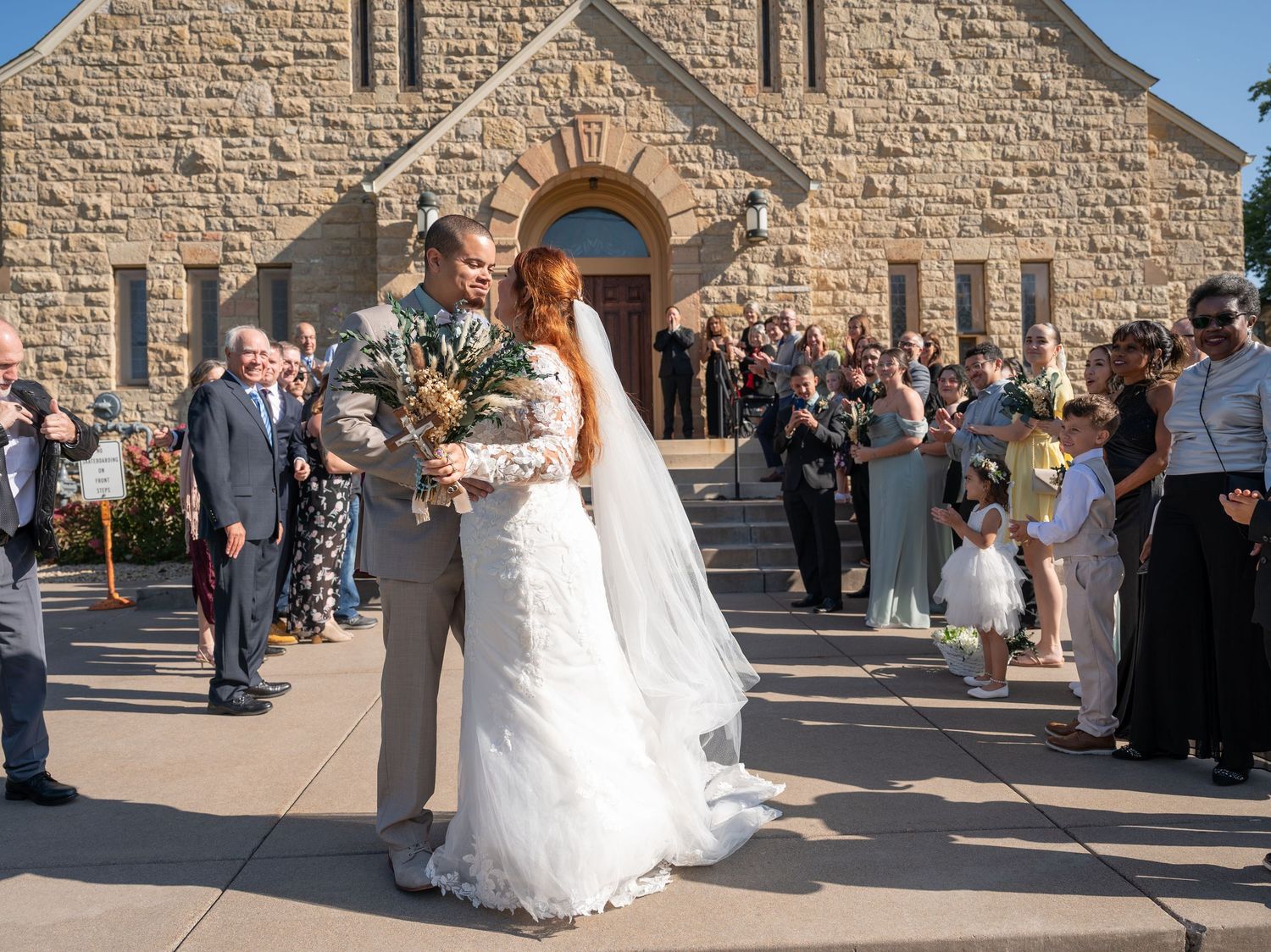 Bride and groom share a kiss outside a stone church surrounded by wedding guests on a sunny day.
