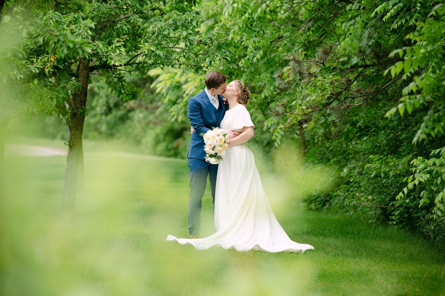 A romantic wedding couple embraces in a lush green garden setting framed by soft focus foliage.