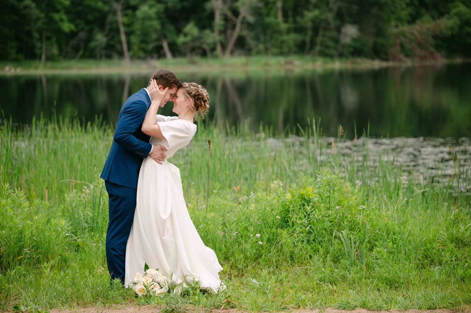 Wedding couple embracing by a serene lake surrounded by lush greenery.