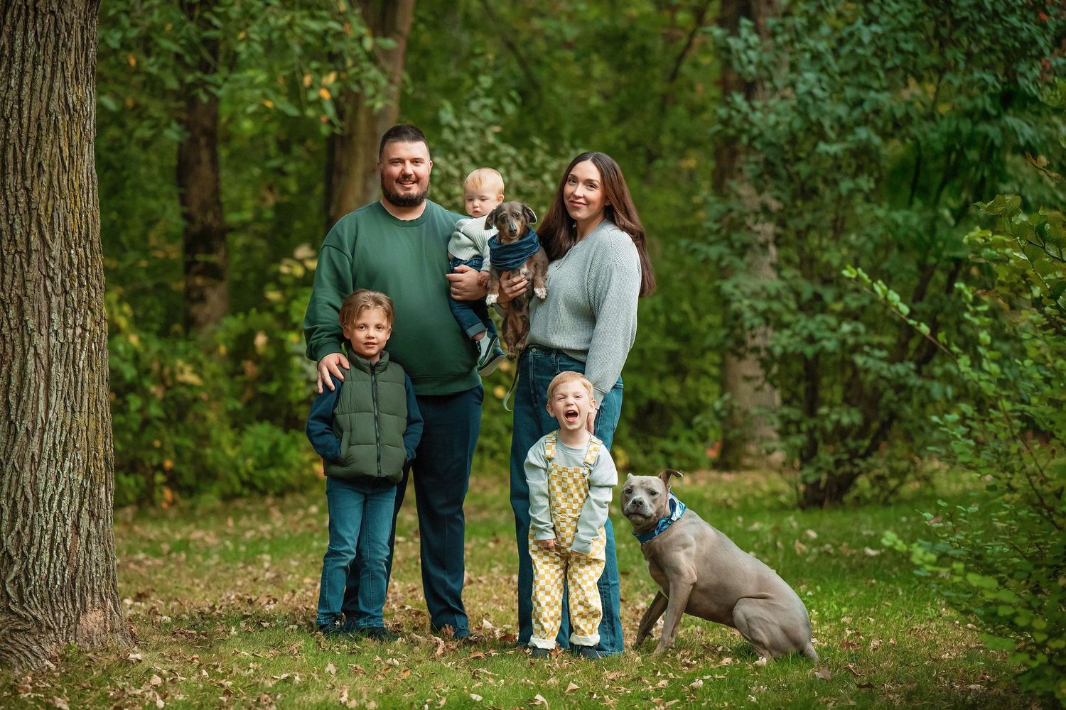 A group enjoys a casual outdoor photo session in a wooded park setting with their dog on a fall day.