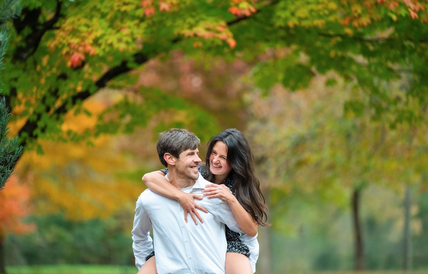 A couple shares romantic moments in a lush autumn park with vibrant fall foliage in the background.