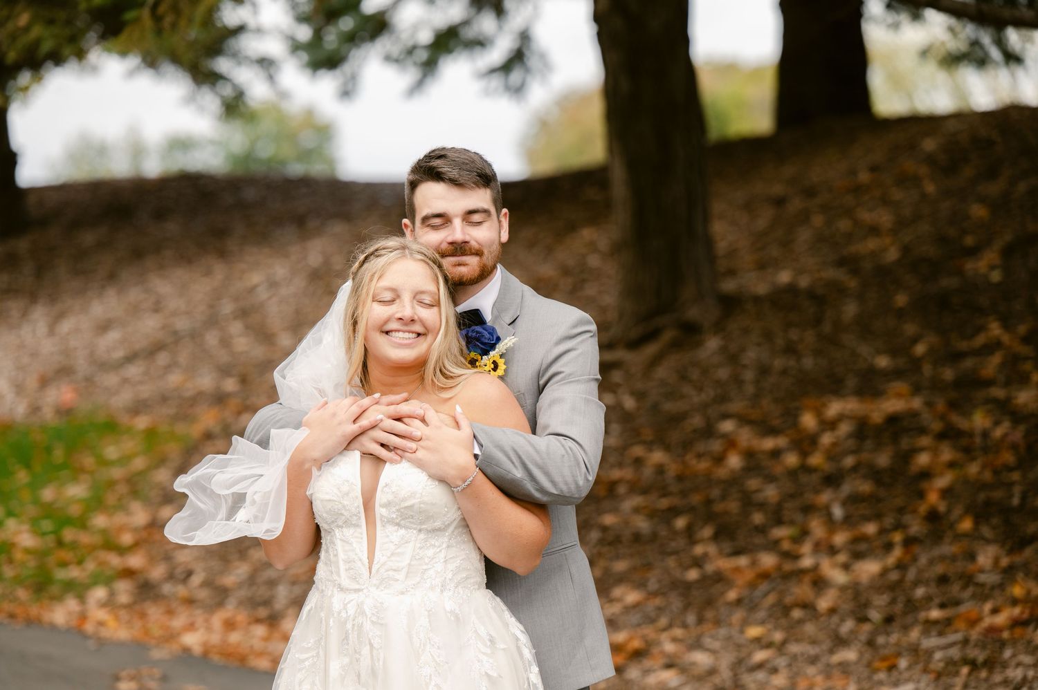 Central MN Wedding Photos | Bride and groom share a joyful embrace on a leaf-covered path surrounded by autumn trees.