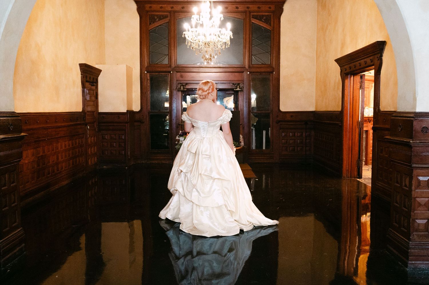Central MN Wedding Photos | Bride in voluminous white ballgown standing alone in elegant dark wood ballroom with chandelier.