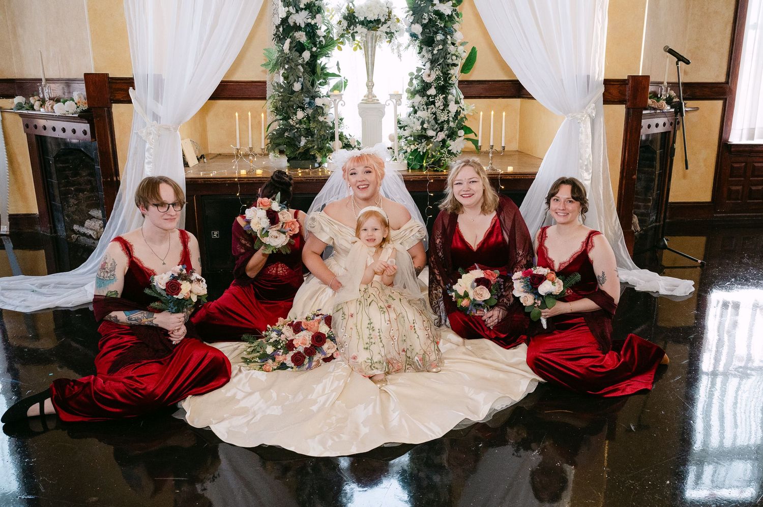 Central MN Wedding Photos | Bride in ivory ballgown poses with four bridesmaids in red velvet dresses and flower girl on marble floor.