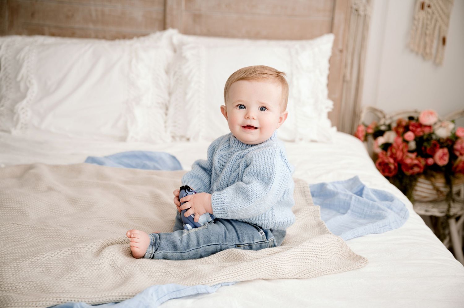 Central MN Milestone Photography - A cute baby in a blue knit sweater sits on a bed with a floral arrangement nearby.
