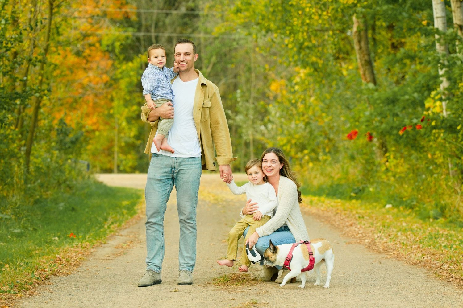 Family with two young children and a dog posing on a scenic autumn trail surrounded by colorful fall foliage.